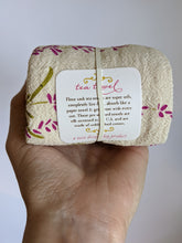 Load image into Gallery viewer, Tea towel rolled up, held in hand. Towel has green and purple lavender print on unbleached cotton. Tag shows it is 100% cotton, lint free, made in USA. Perfect for spring!