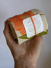 Load image into Gallery viewer, Orange and green printed poppy flower shown on rolled up tea towel in hand. Tag on front tells you the flour sack tea towels are super soft, lint free, 100% cotton, made in USA.