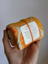 Load image into Gallery viewer, Rolled up tea towel with oranges print shown held in hand with tag showing these are flour sack, 100% cotton. 
