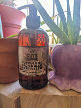 Cargar imagen en el visor de la galería, Succulent, Cacti, &amp; Aloe plant fertilizer amber colored pump container shown next to aloe plants on a sunny shelf to show it looks nice sitting out. No need to put it away like some other brands that aren&#39;t as pretty. 
