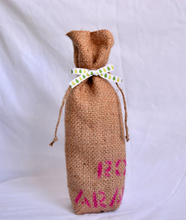 Cargar imagen en el visor de la galería, Burlap wine bag with pink text on the bottom half.  This is made from recycled coffee bean sack. The text originally told the origin of the coffee . It ties shut with a festive ribbon. 
