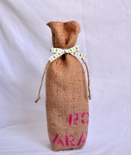 Burlap wine bag with pink text on the bottom half.  This is made from recycled coffee bean sack. The text originally told the origin of the coffee . It ties shut with a festive ribbon. 