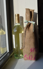 Cargar imagen en el visor de la galería, Burlap wine bag next to wine bottle to show scale of size. Bag is made from recycled coffee bean sack with pink text visible at the bottom half of it. Pink text originally told origin of coffee. 
